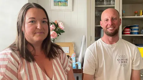 Sarah Doolin and Craig Pollard are smiling as they sit in their kitchen. Ms Doolin is wearing a terracotta and white striped blouse and has long brown hair. Mr Pollard has a shaved head and brown beard and is wearing a white T-shirt with a Volcom logo. Behind them is a shelving unit with glasses and mugs lined up and there is a vase of flowers. On the wall there is a landscape picture.