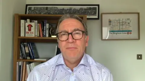 Kieran Maguire is wearing glasses and a white spotted shirt and looking directly at the camera. Behind him is a book shelf and a frame photo of The Brighton Line