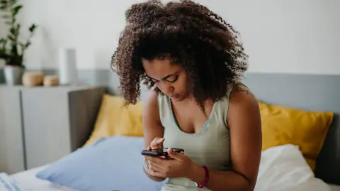 A young woman sits on a bed looking down at her phone screen.