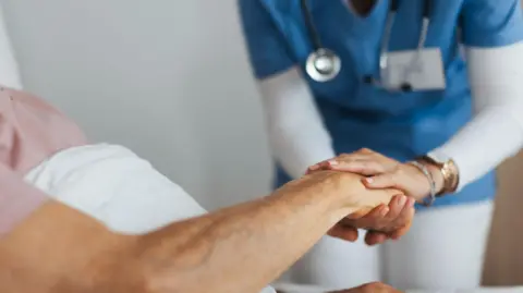 A caregiver holds the hand of a senior client who is in bed. 