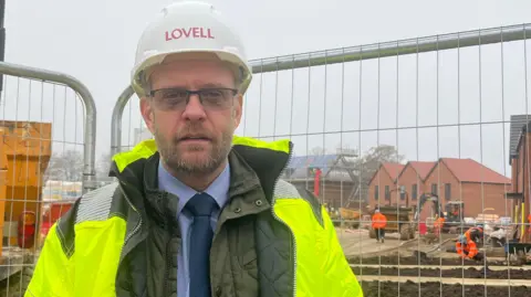 A man wearing a white hard hat, glasses, and a hi-vis jacket over a shirt and tie. He is standing in front of the metal fencing around a building site while looking into the camera