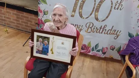 Betty sitting on a chair holding a framed photo of her letter from the King and Queen. She sits in front of a big sign saying happy 100th Birthday. She looks happy.