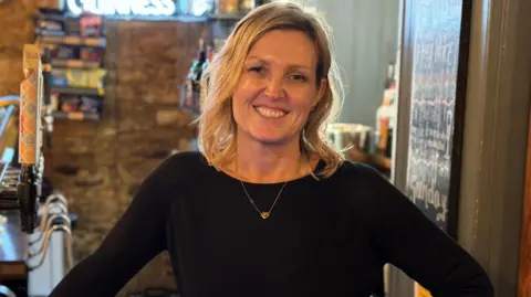 A blonde woman smiling at the camera. She is standing behind the bar in a pub. She is wearing a black top with a gold necklace.