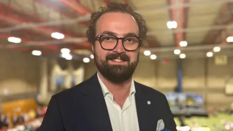 Head and shoulders image of Ross Martin in a sports hall. He has short curly dark hair, rounded dark glasses and a beard. He is wearing a dark suit jacket and white shirt.