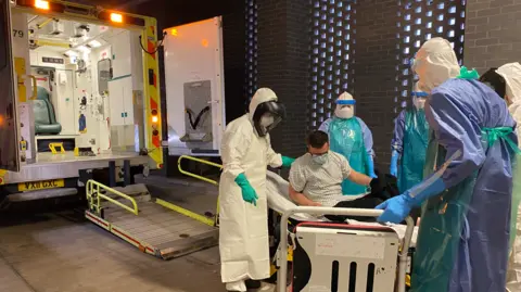 A simulation exercise at Southmead Treatment Centre for patients with dangerous airborne infectious diseases. Five staff are in white boiler type suits, wearing full PPE and standing around a patient on a gurney. They are loading them into an ambulance. 
