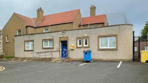 In the back ground is a two-storey building. In the foreground is a single-storey grey building with flat roof.