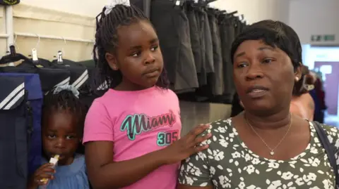 A mum wearing a olive green and white dress in a floral pattern standing in a shop full of clothing on racks with her two girls next to her - one is in a pink T-shirt and the other is in a blue summer dress. 