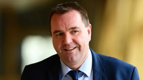 A man with dark hair smiles while walking in the Scottish Parliament. He is wearing a dark blue suit and tie, with a light blue shirt. He is visible from the shoulders up. 