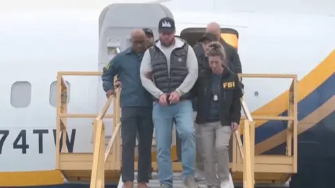 AFP Ryan Wedding verlaat een vliegtuig met zijn handen in handboeien. Wedding draagt ​​een spijkerbroek, een zwart vest en een zwarte baseballpet. Hij wordt geflankeerd door twee FBI-agenten: een man en een vrouw. 