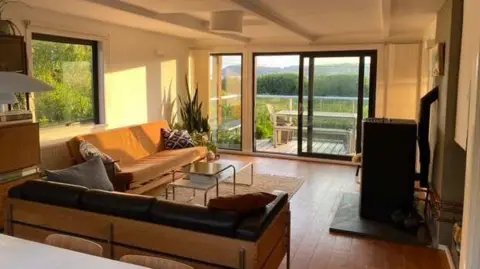 A large living room with 60s style sofas in an L shape, facing a wood burner. The sun is beaming in through the open French doors overlooking the estuary.