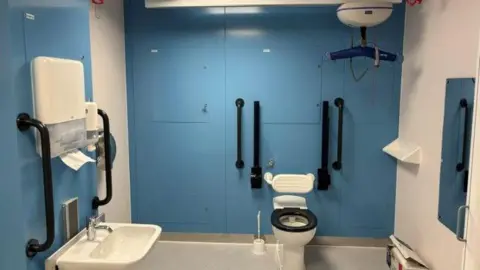 North West Anglia Foundation Trust A changing room with a white floor and blue walls, a white and black toilet, a blue and white hoist on the ceiling, a white bin and sink.
