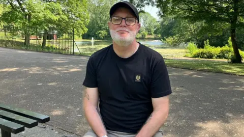 A man sat in a park looking towards the camera. He is next to a dark green bench and is wearing a black t-shirt and a cap. He has glasses and a beard. 