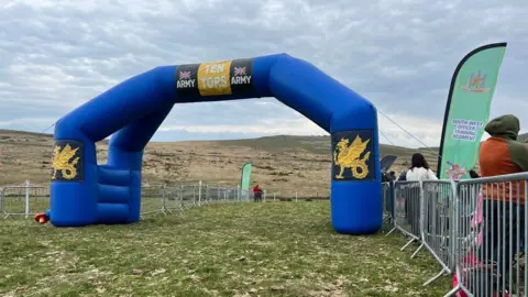 The finish line at the Ten Tors Challenge. It is an inflatable blue archway with the words Ten Tors written on it. There are people waiting next to a metal barrier fence near the finish line.