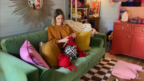 A woman sat on a green sofa knitting. She has long hair and is wearing a gold knitted top. She is using red wool