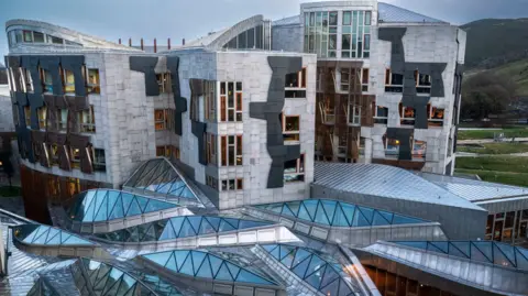 The Scottish Parliament building. It is made of concrete and coloured blocks and is standing in front of a hill.