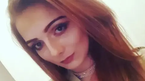 Facebook A woman with long ginger hair poses in as selfie as she wears makeup, a light brown colour top and sparkly choker necklace.