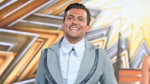 Getty Images Paul Danan wearing a blue striped shirt with a large collar underneath a grey blazer. He is smiling and looking upwards as he walks across a stage decorated with gold star patterns.