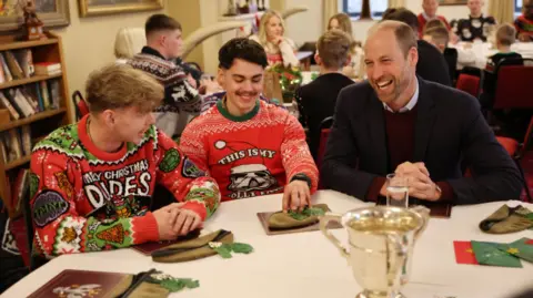 Richard Pohle/The Times/PA Wire Prince William sitting at a table with two young military men wearing Christmas jumpers and smiling, in a room with lots of other families 