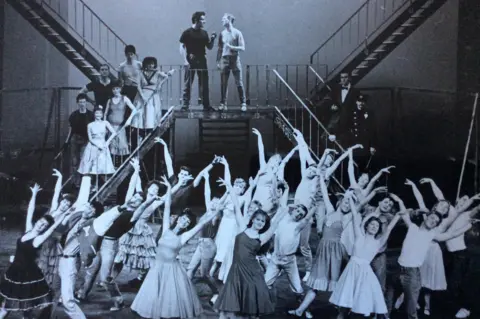 Northern Theatre More than two dozen men and women dressed in 1950s outfits dance on a stage set. Most of them are at the front with their arms raised high in the air. Others stand among four large flights of steps, which are arranged in an X shape.