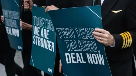 PA Pilots on strike holding signs that read 'together we are stronger' and 'two years talking, deal now'. The pilots are in uniform (black blazers and trousers with yellow stripes on the cuff and a plane gold badge on the lapel.) The signs are green/blue with white writing.