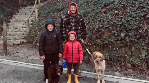 A man in a checked jacket and red hood with two boys in black and red coats and a spaniel and labrador