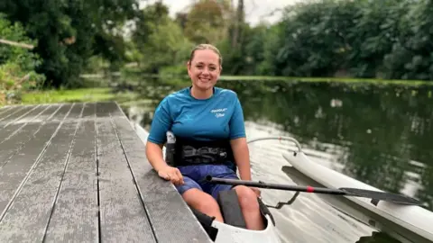 Saffron Cresswell A woman with her hair tied back, wearing a blue T-shirt and shorts, strapped into an adapted canoe on a river, with trees and banks behind her and wooden decking to her left.