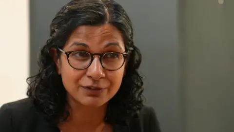A woman with curly dark hair and black rimmed glasses. She wears a black blazer and a polka dot dress or top. She looks to be of South Asian heritage.