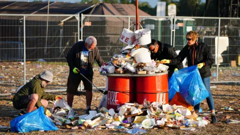 PA A group of people picking up litter that surrounds overflowing red bins at Glastonbury and collecting it in blue plastic bin bags. The ground is muddy and covered in litter and food packaging.