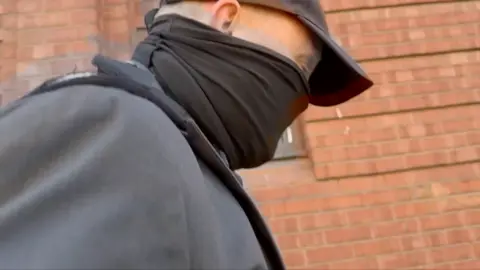 Robert Bush wearing a black cap, with the bottom of his face covered up.