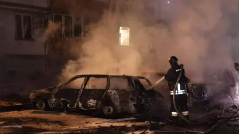 Office of Ukraine Attorney General/Telegram A firefighter firing water at a burnt our car, smoke surrounding them.