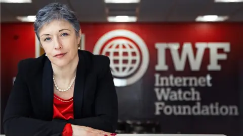 IWF Sisue Hargreaves sitting at a desk with her organisation's name - the Internet Watch Foundation - decorating a wall behind her