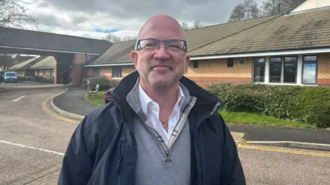 Ed Wilson stood in front of a brick building. He is wearing a dark jacket, grey jumper and a white shirt. He is a bald man with blue glasses. 