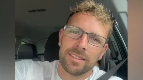 A man with curly blonde hair is wearing glasses and a white t-shirt while sitting in a car. He is looking at the camera.