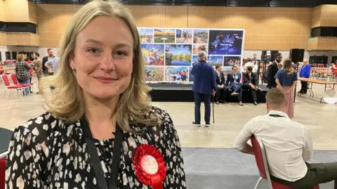 Amy Holmes/BBC Lauren Townsend smiles at the camera wearing a red Labour rosette. She stands with the election hall behind her, including a colourful montage of images from across Milton Keynes.