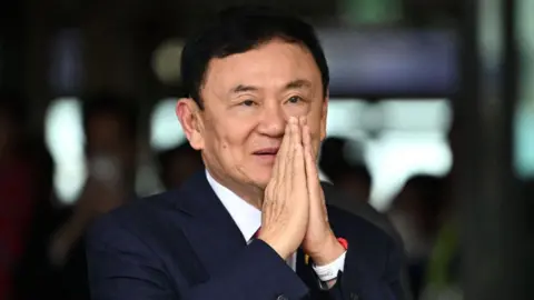 Close-up of former Thai prime minister Thaksin Shinawatra in a suit, palms together together, against a blurred background.