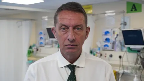 A middle-aged man faces the camera wearing a white shirt and black tie with the equipment of a hospital ward seen behind him