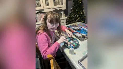 Eileen Mitchell A teenager is sitting at a kitchen table doing arts and crafts. She is wearing a pink top and has long brown hair. There is a bowl of coloured rocks and glue next to her. A Thomas the Tank Engine mat is lying on the table. A Christmas tree is in the background. 