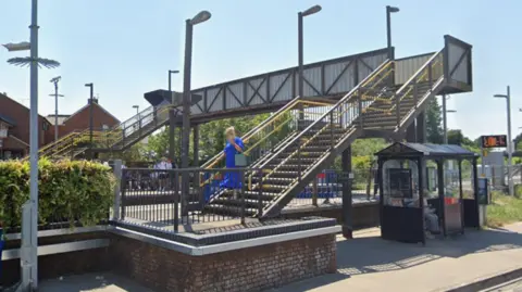 Google Street view image shows the footbridge crossing the railway line at Dorchester South Station. The metal bridge, viewed from the nearby road, has steps up from the platforms.