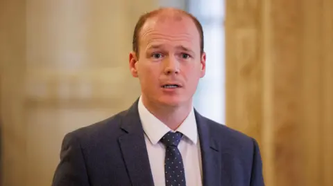 PA Media Lyons is wearing a navy suit with a white shirt and a navy tie with pink spots. He's talking. The background is out of focus. 