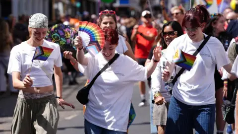 PA Media Three people wearing white T-shirts wave LGBTQ+ flags and fans.