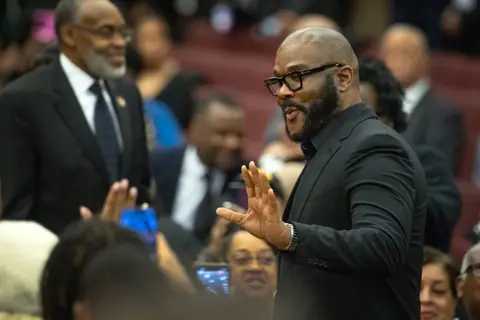 Getty Images Filmmaker Tyler Perry is seen standing at the service wearing a black suit and black eye glasses.