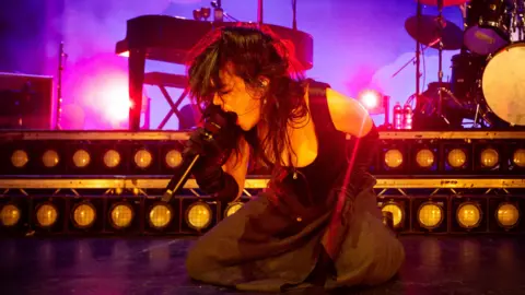 Getty Images Abigail Morris of The Last Dinner Party performs at O2 Academy Leeds on 24 September. Abigail kneels on stage as she sings into a microphone, her long dark hair worn loose. She wears a black skirt, long black gloves and corset-style top. Behind her there's a piano and drum kit, the stage is lit purple behind her. 