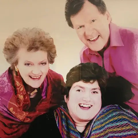 Rachael Scott-Hunter A family portrait. Alexandra has short brown hair and wears a black top and multi-coloured shawl. Rachael has short grey hair and wears a dark top and a red/pink shawl. Ian has short dark hair and wears a smart pink shirt. All are smiling broadly.