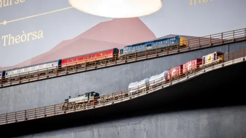 Paul Campbell The tracks for the model railway run along the walls of the Victorian Market. On the lower track a steam locomotive hauls a freight train. On the track abaove a blue and yellow diesel locomotive pulls red and blue carriages.
