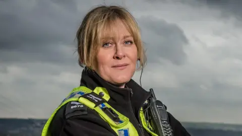 BBC/Red Productions Sarah Lancashire as Catherine in Happy Valley. She is wearing a police uniform and standing against a background of hills are grey skies.