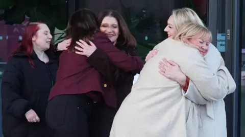 PA Media Two pairs of women are hugging, with a fifth watching. The pair nearest are both wearing cream coats and have blond hair. Next to them are two wearing maroon coats and with dark hair. The woman standing nearby has dyed red hair and is wearing black. They are all beaming with delighted smiles.