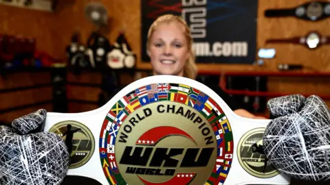 John Fairhall/BBC Turner is seen holding her WKU K1 World Champion belt. She is smiling while holding the white belt with a gold plate detailing the title.