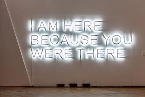 Richard Ivey Neon words displaying the text I AM HERE BECAUSE YOU WERE THERE are displayed on a white wall with a laminate floor in the foreground