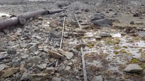 Tim Osborne Corroded rail tracks on a rocky beach next to a rusted and broken iron outlet pipe at low tide.