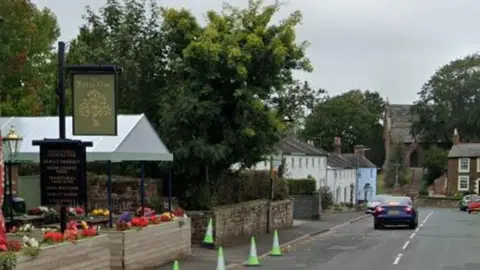 Royal Oak Pub on left with a green and yellow sign reading Royal Oak and flower boxes. Green cones line the road down the village. A church can be seen in the distance. 